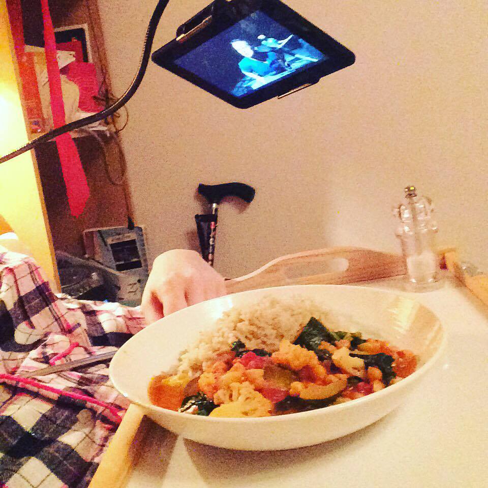 A sideways view of someone eating in bed, with a tablet held by a tripod overhead. You can see their food in a bowl.