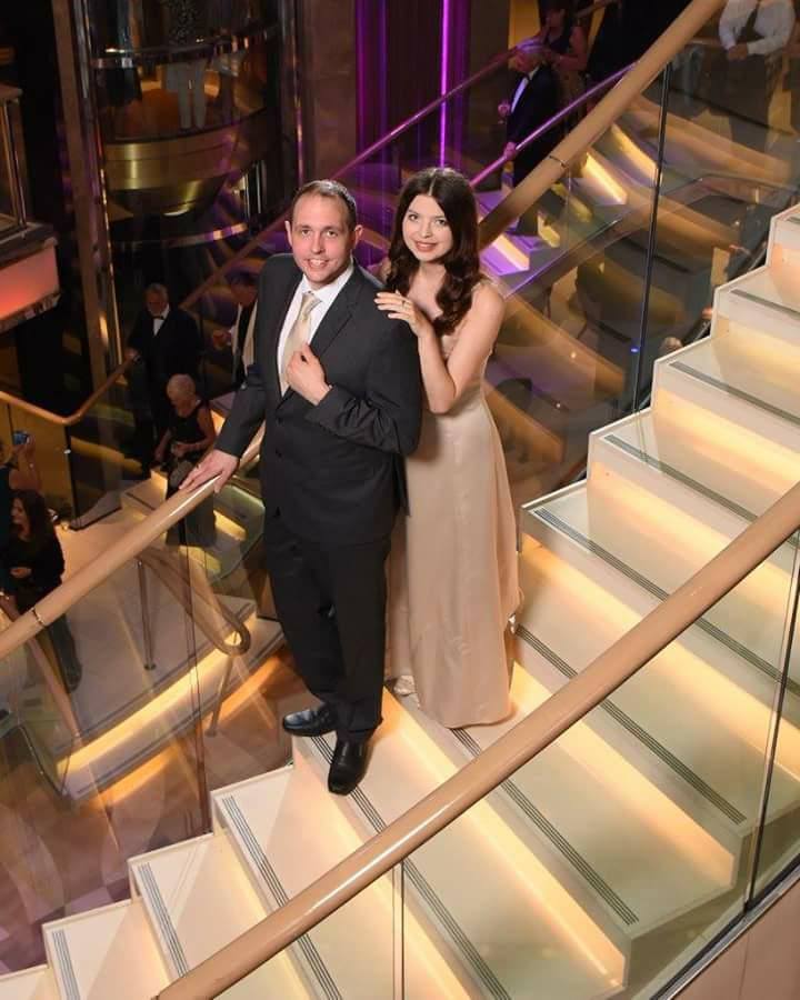 Jenni and a gentleman wearing evening outfits, standing on a lit staircase.