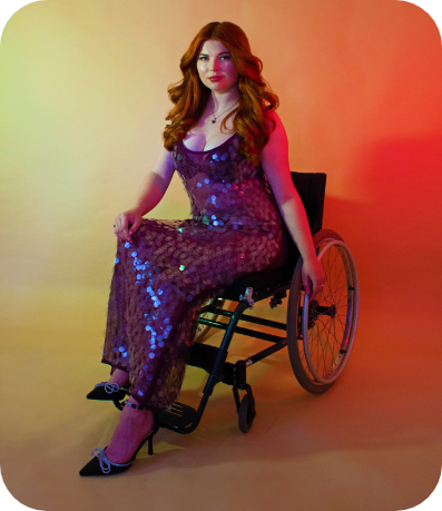 A professional modelling shot of Jenni sat in her wheelchair, wearing a dazzling giant sequin purple iridescent dress. She is sat against a light yellow/orange ombre background.