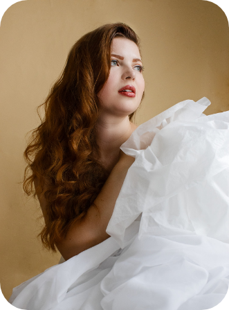 Jenni looking to the right, covered in soft white fabric. Her long auburn hair trails her shoulder with soft curls, complementing her neutral, rosy makeup.