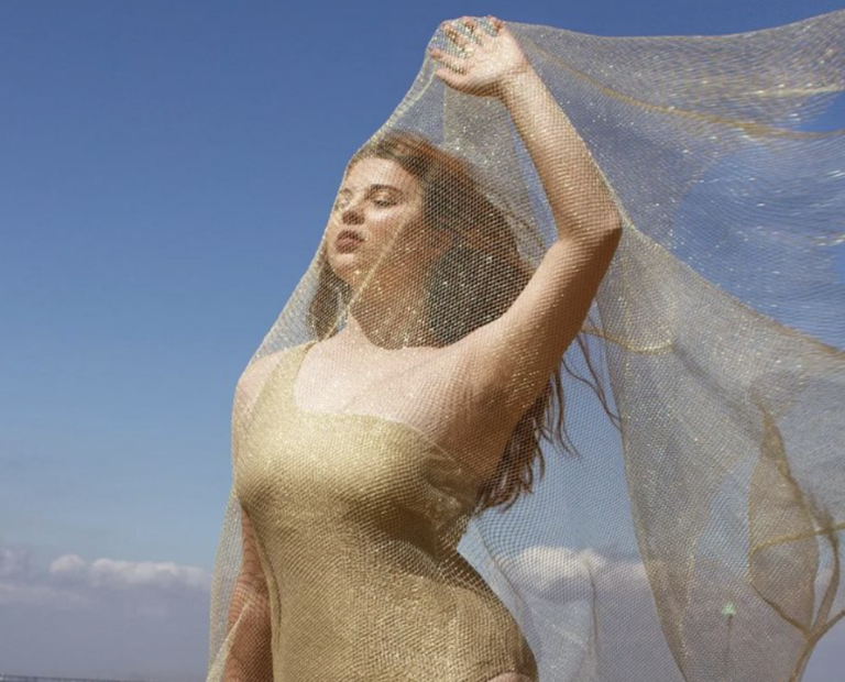 A professional shot of Jenni modelling a gold swimsuit against a blue sky. She is holding a mesh material over her face and has her eyes shut,facing the sun. Her makeup is gold to complement the outfit.