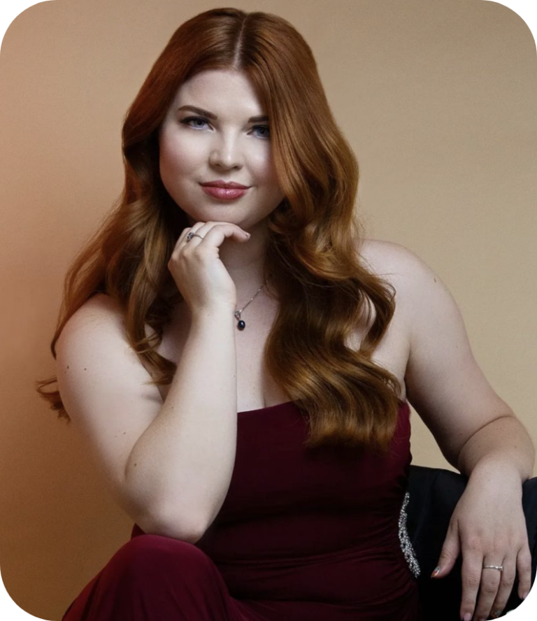 A professional model shot of Jenni looking at the camera and slightly smiling. She has long auburn hair which is curled and is wearing a red dress.