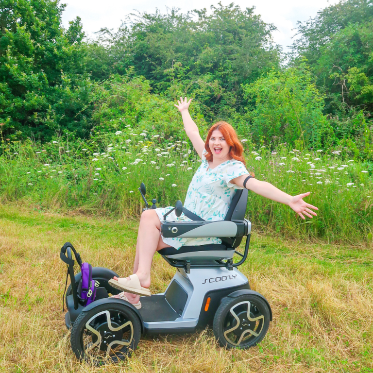Jenni in her mobility scooter Scooty in a grassy field, with her arms out, smiling.