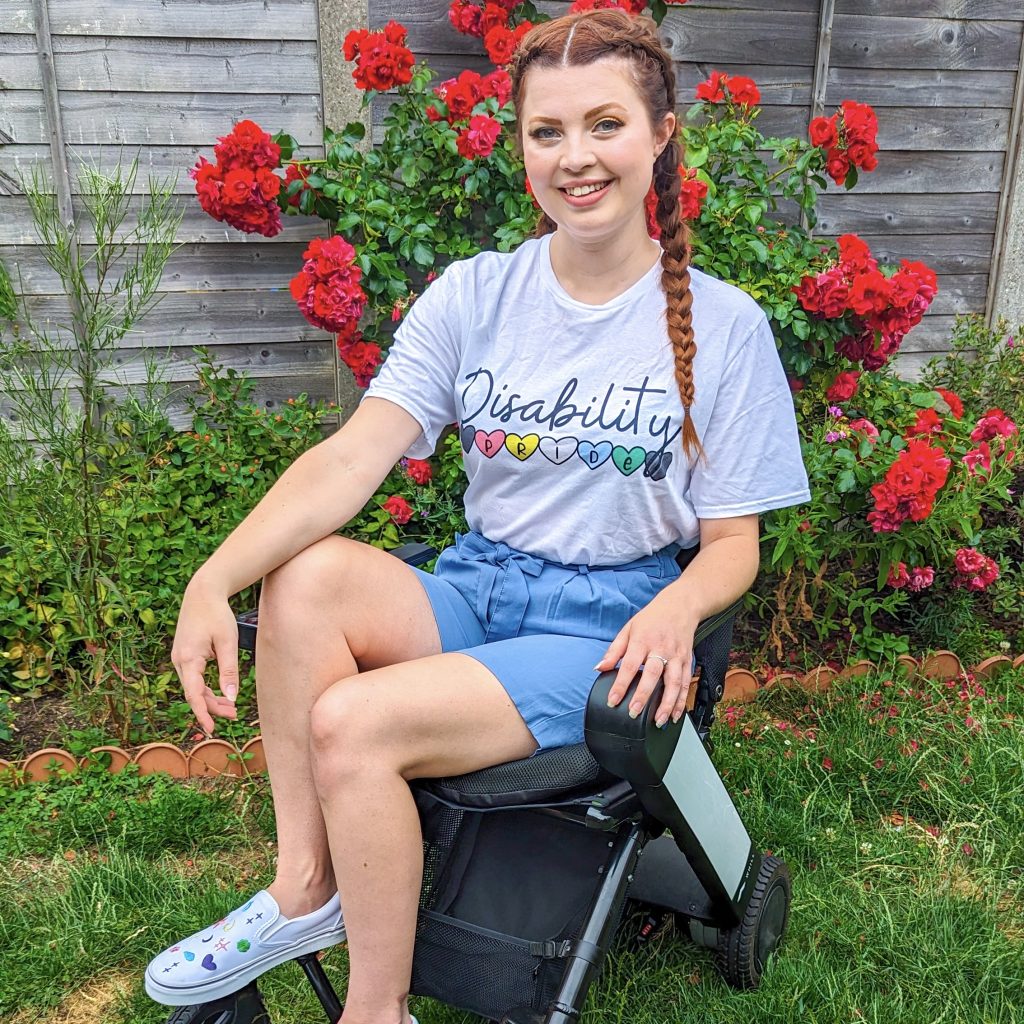 Jenni sitting in her garden, in her wheelchair, wearing a 'disability pride' t-shirt and blue shorts, smiling.