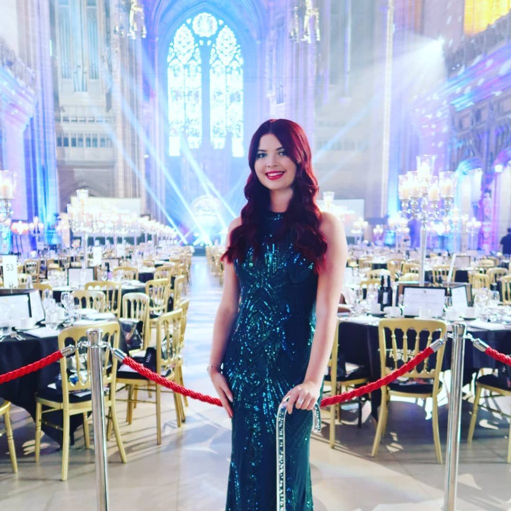 Jenni stood in front of an event with lots of seating, wearing a green beaded dress. She is smiling and leaning on her stick.