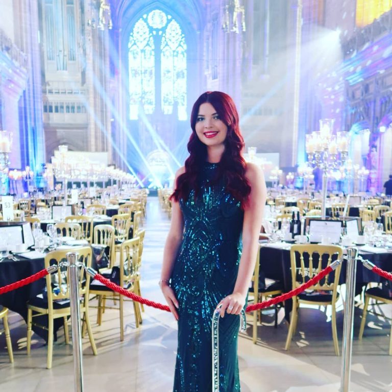 Jenni stood in front of an event with lots of seating, wearing a green beaded dress. She is smiling and leaning on her stick.