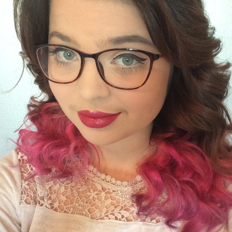 A close-up photo of Jennis face, with her brown to pink ombre hair in view. She is wearing a white lace top, and has glasses.