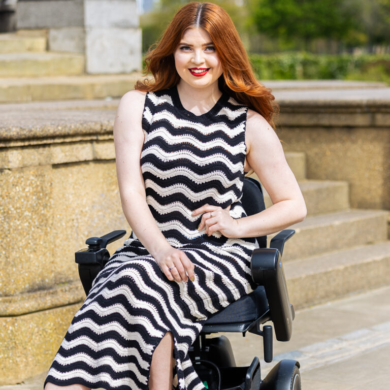 Jenni sitting in her wheelchair, looking towards the camera smiling. She is wearing a black and white chevron dress with black heels.