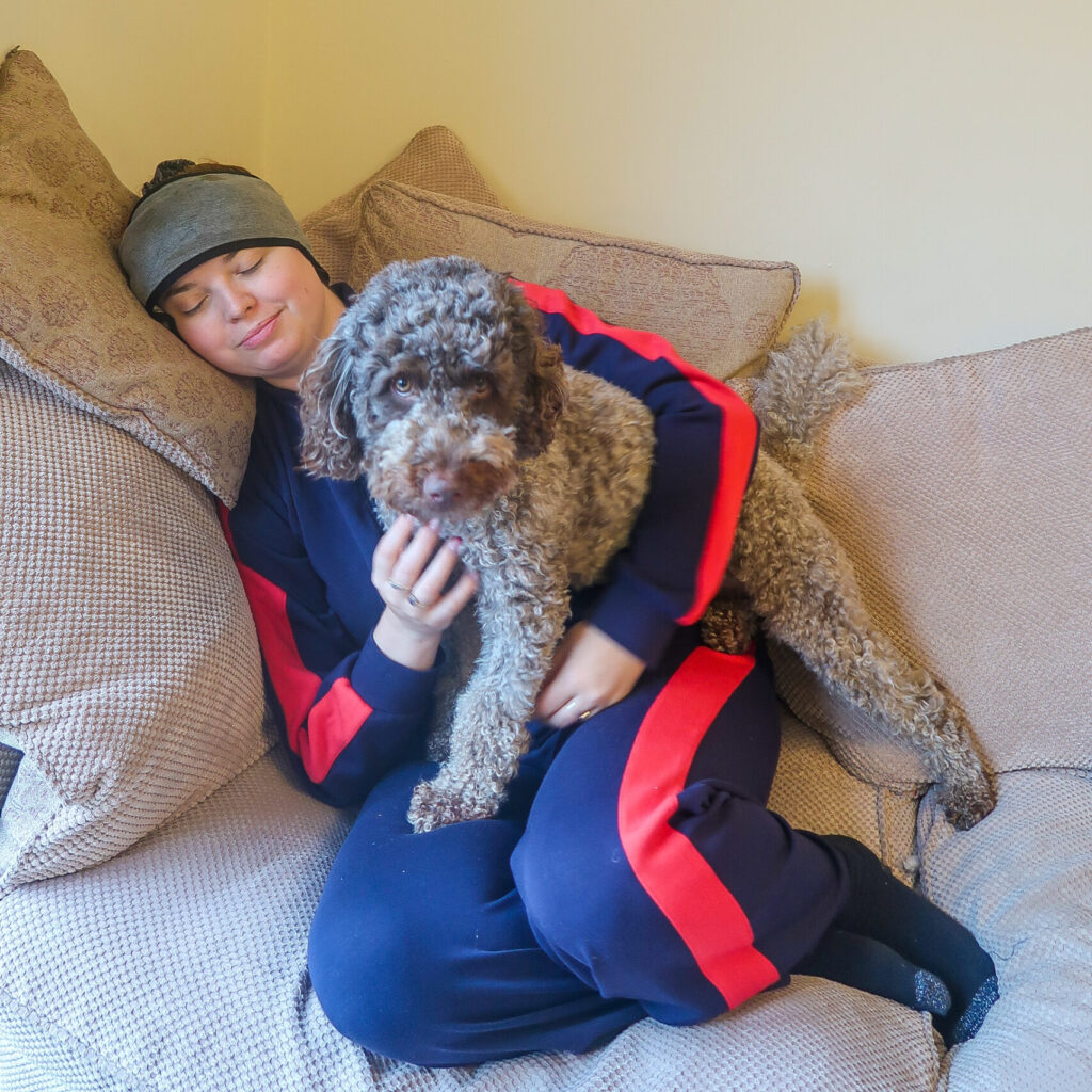 Jenni lying on the sofa managing migraines by wearing a headache headband with her eyes closed. Her labradoodle dog Coco is sat on her lap looking worried about her.