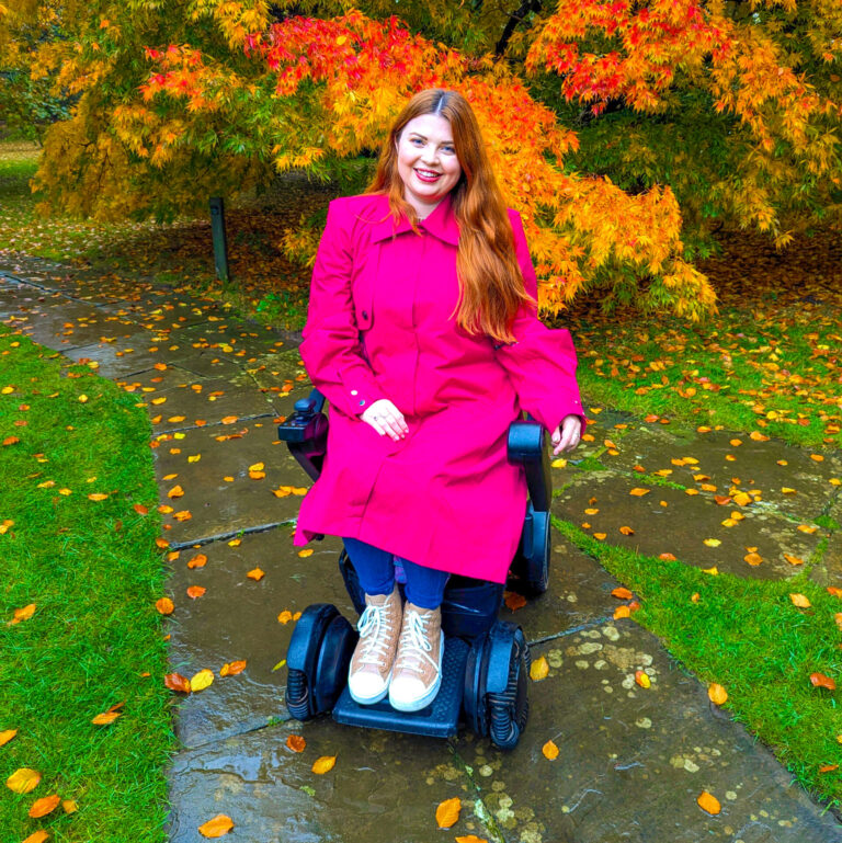 Jenni sat in an autumnal garden in her electric wheelchair wearing accessible clothing which is bright pink adaptive coat