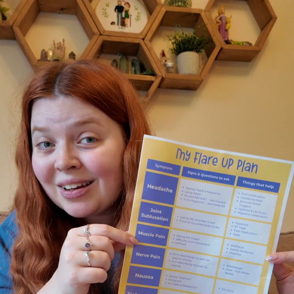 A woman with long red hair, wearing a blue top, smiles while holding up a printed “My Flare Up Plan” chart. The chart has a yellow and purple color scheme and lists symptoms like headache, joint subluxation, muscle pain, nerve pain, and nausea, along with signs, questions to ask, and things that help. Behind her is a decorative wall with honeycomb-shaped shelves containing plants, ornaments, and small figurines.