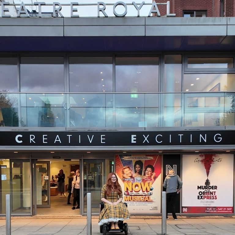 Jenni a white woman with auburn hair and glasses is sat in her wheelchair outside Norwich Theatre Royal. She is wearing a yellow 50s style dress and there is a poster for murder on the orient express behind her.