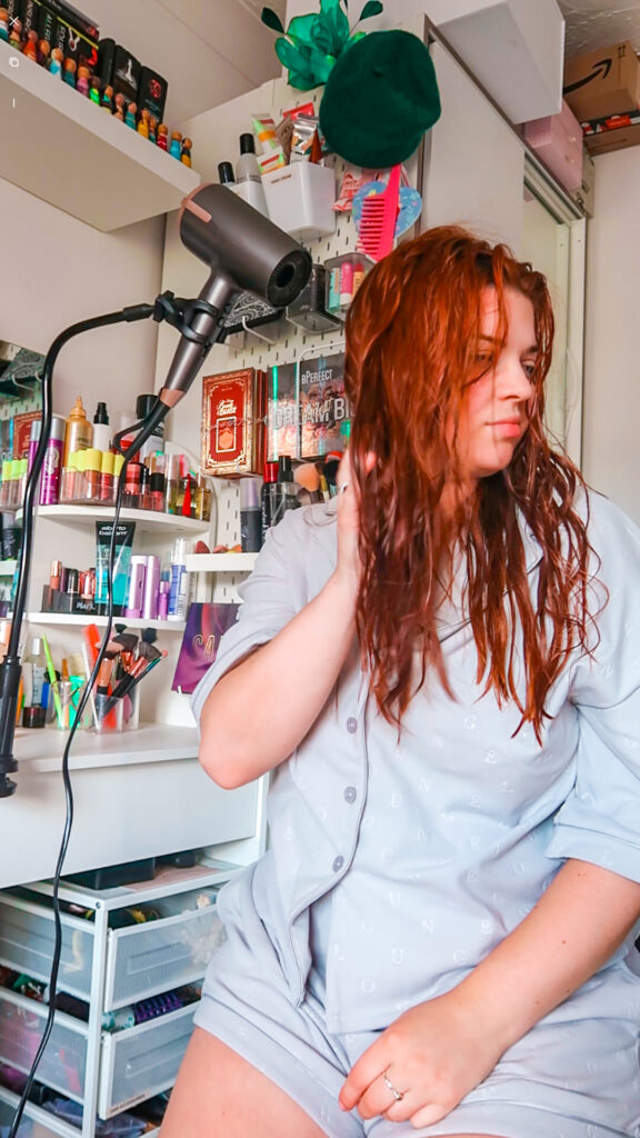 Jenni is sat at her dressing table with half wet hair. A black hair dryer stand is clipped to her dressing table at one end and holding up a grey hairdryer at the other, drying Jenni's hair