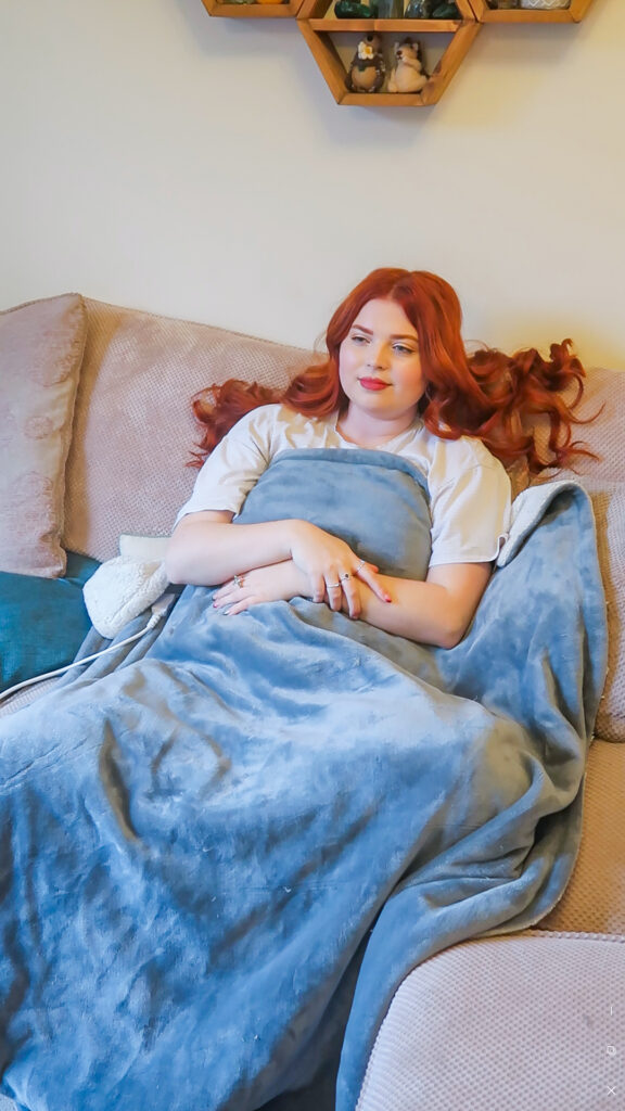 Jenni is sat on her beige sofa wrapped in a grey electric heated throw blanket with a fluffy white underside