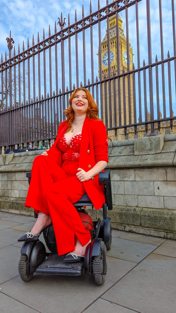 Jenni sitting in her wheelchair in a red suit, with a lacy top in front of Big Ben in London.