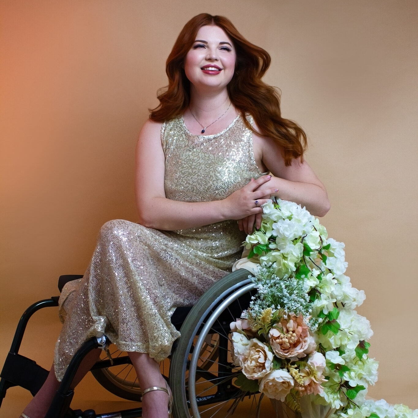 Jenni sat in her wheelchair, with flowers trailing off of it. She is wearing a gold sequin dress with heels.