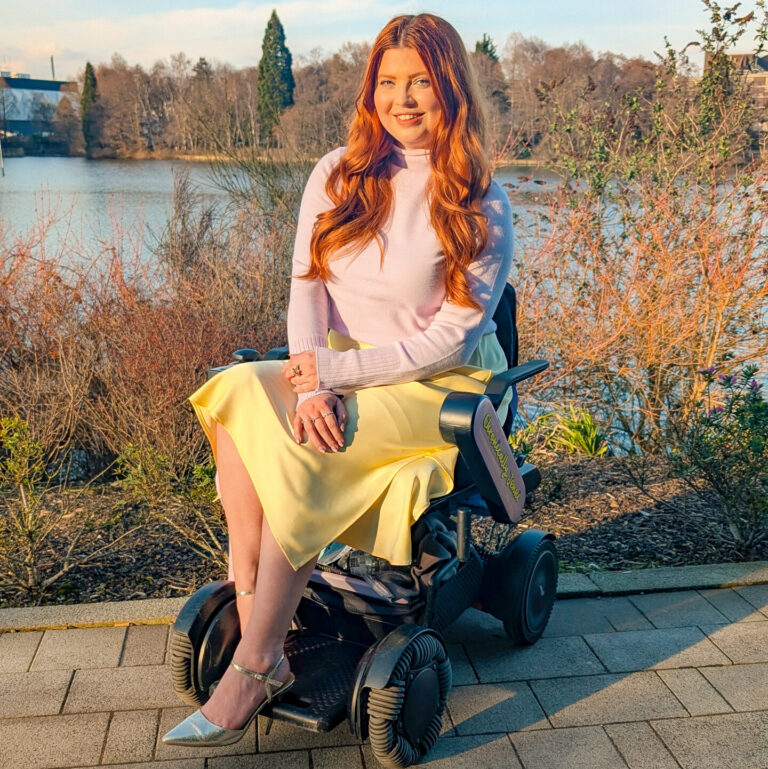 Jenni a white disabled woman with wavy auburn hair. She is sitting in her powered wheelchair in front of lake. shes wearing a lilac turtle neck with a pastel yellow silk skirt and gold heels