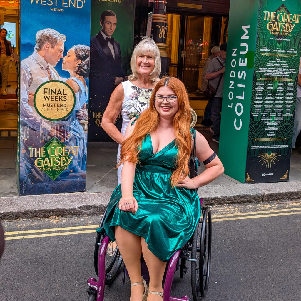 Jenni and her mum posing outside the london coliseum with the great gatsby banners in the background. Jenni is sitting in her manual wheelchair wearing a green sparkly dress and her mum is wearing a cream floral dress.