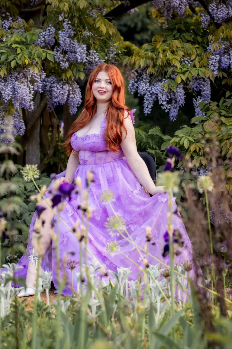 Jenni standing outdoors beneath cascading wisteria blossoms, smiling warmly while wearing a floral dress.