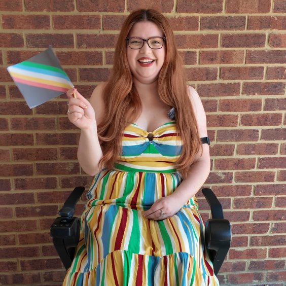 Jenni, a smiling white woman with long red hair and glasses, sits in a power wheelchair in front of a brick wall. She wears a colourful striped dress in red, yellow, blue, and green. She is holding a small Disability Pride Flag with five horizontal stripes in green, blue, white, yellow and red on a black background.