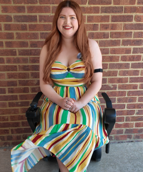 Jenni, a smiling white woman with long red hair, sits in a power wheelchair in front of a brick wall. She wears a colourful striped dress in red, yellow, blue and green, with matching green shoes featuring beaded bows. Her hands are folded in her lap, and a breeze lifts part of her skirt.